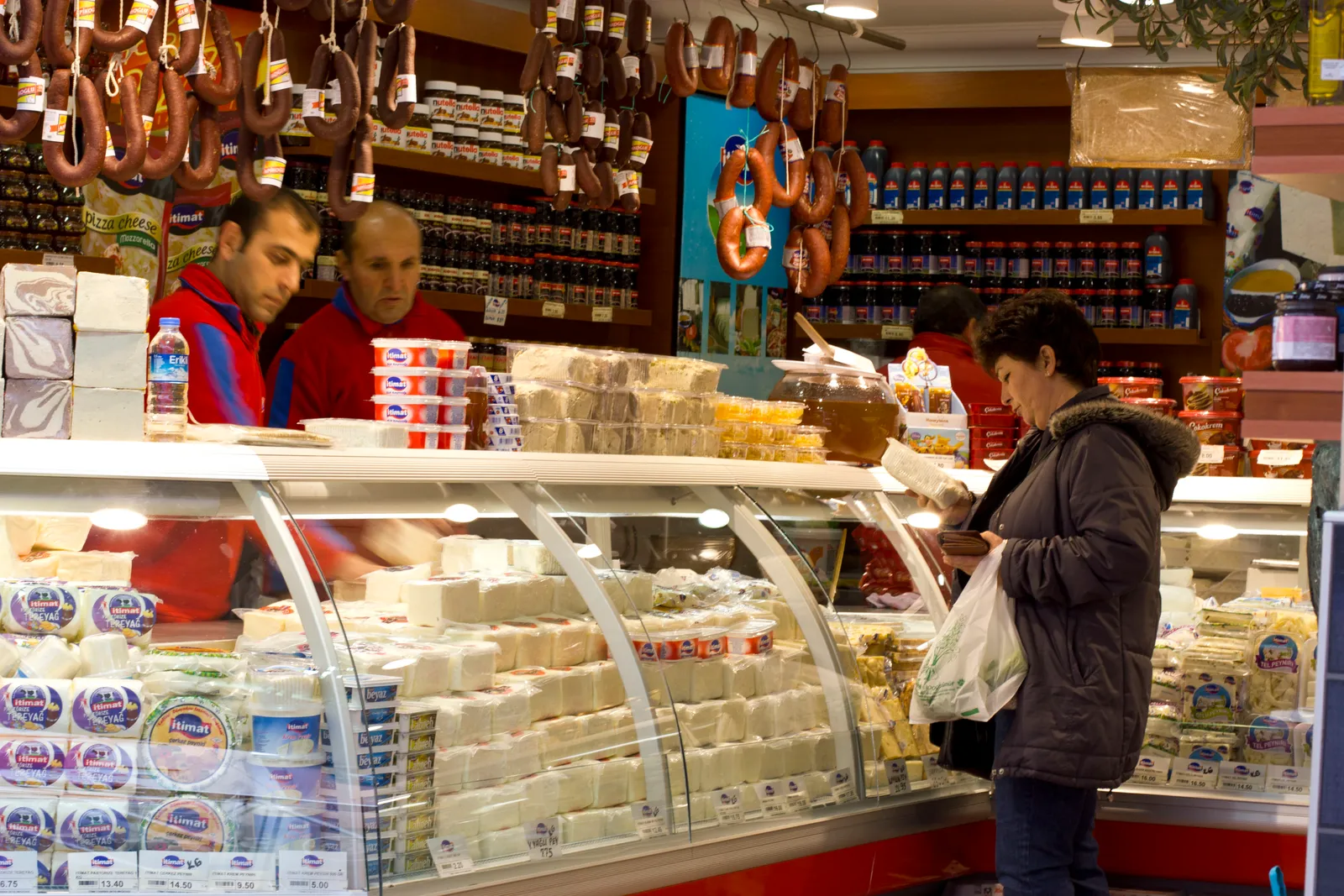 A delicatessen counter in the Kadıköy market in Istanbul, trays of white cheeses under glass, sausages hanging above the till and a customer being served