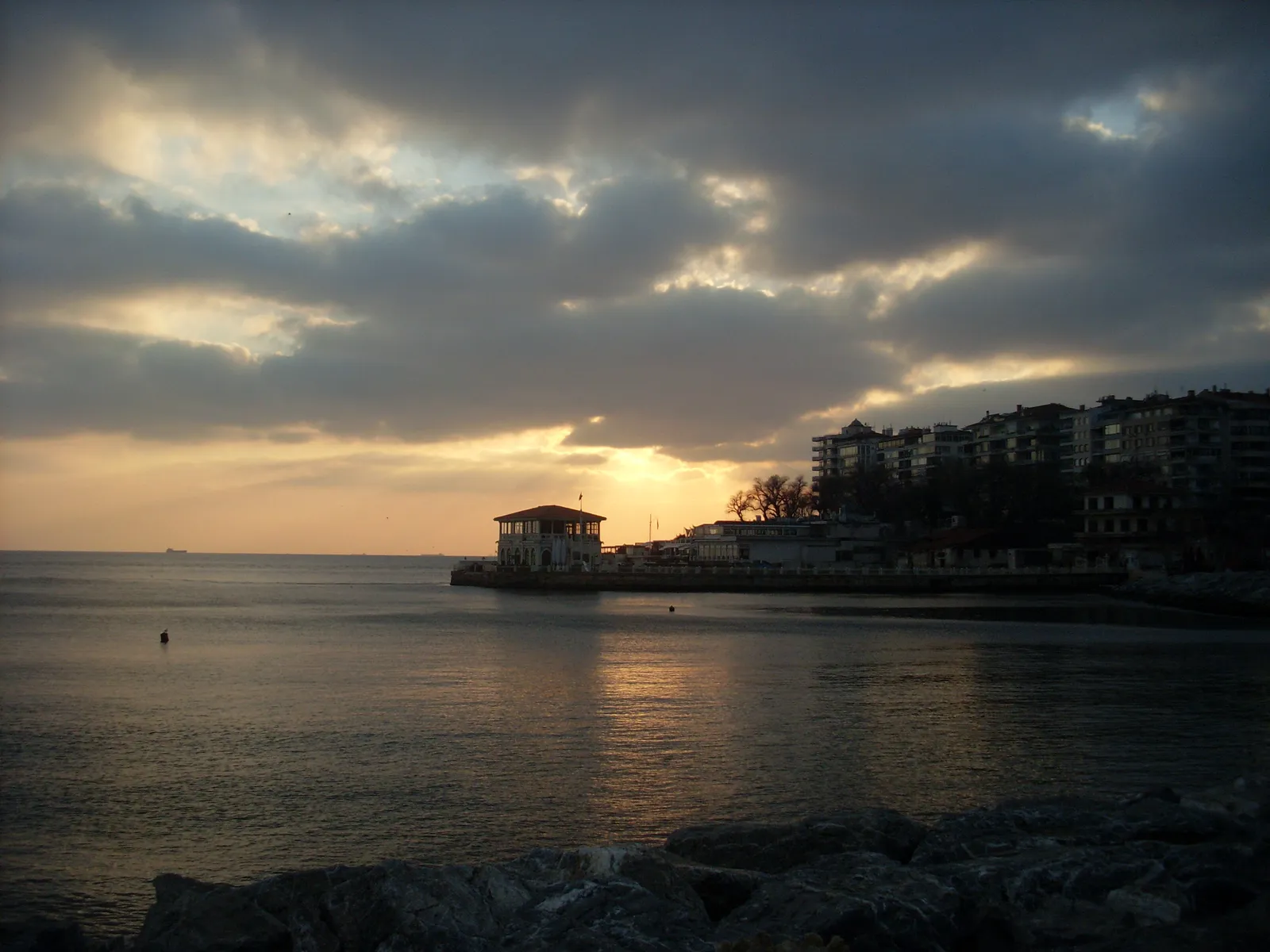 The Moda shoreline in Istanbul at dusk, the Sea of Marmara in the foreground, a small waterfront pavilion silhouetted on the headland and apartment blocks along the shore behind it