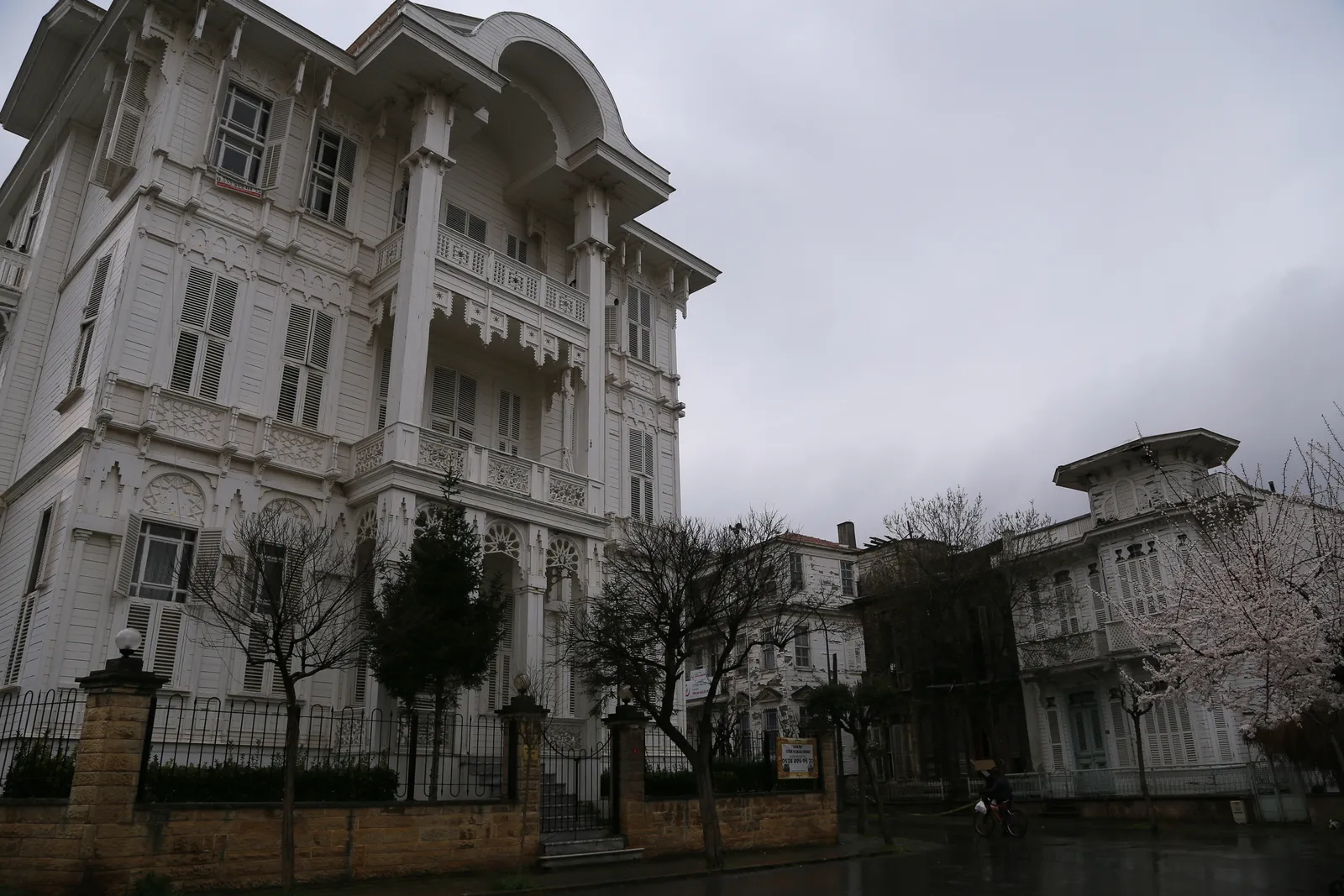A large white 19th-century wooden mansion on Büyükada with tiered balconies and shuttered windows, behind iron railings on a quiet island street with bare trees