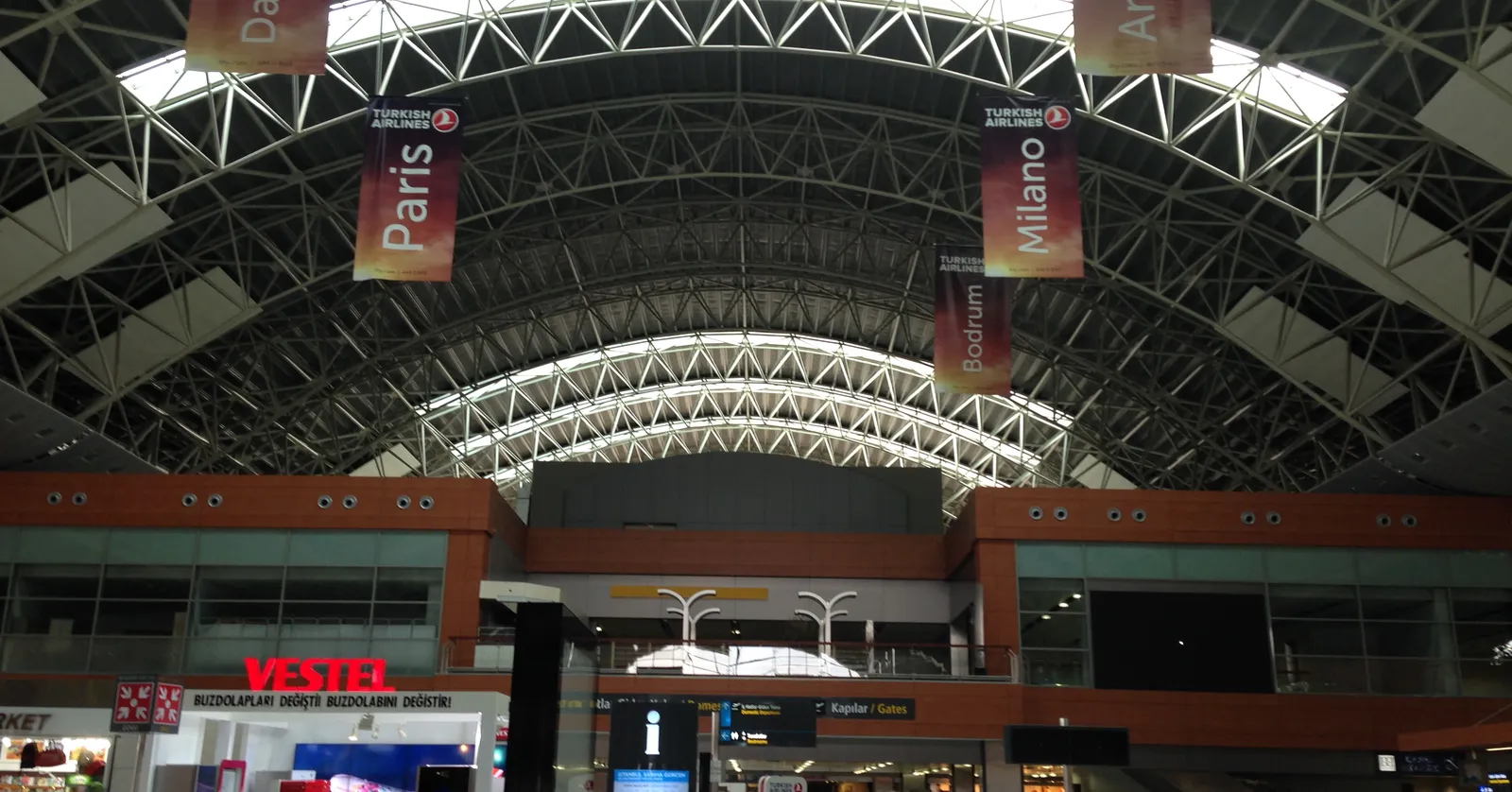 Inside the single terminal at Istanbul Sabiha Gökçen Airport, a long barrel-vaulted steel roof over the concourse with destination banners for Paris and Milan hanging from the trusses