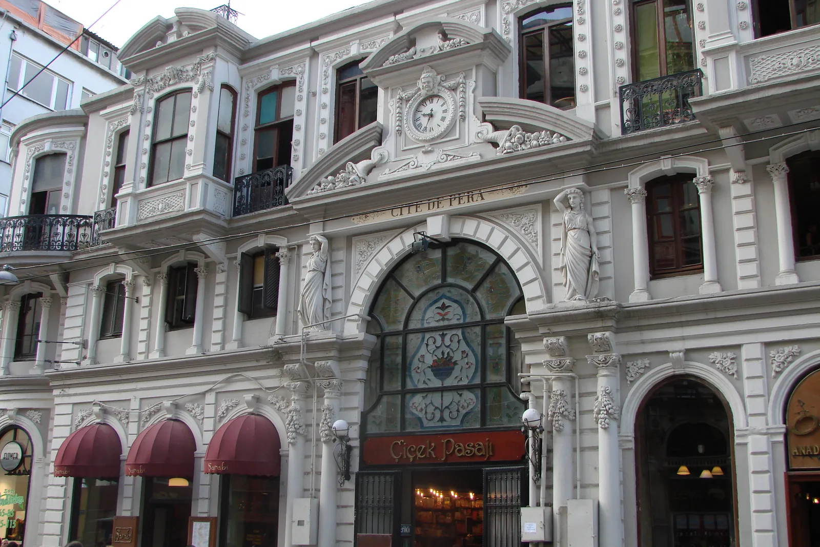 The ornate white facade of Çiçek Pasajı on İstiklal Avenue, with a clock above the central arch, the name Cité de Pera carved across the stonework and restaurant awnings along the ground floor