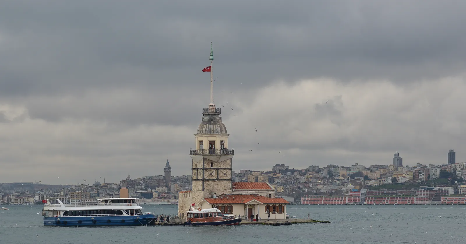 The Maiden's Tower on its islet at the mouth of the Bosphorus, a square white tower with a pointed roof and a Turkish flag above a low red-roofed building, with a passenger boat moored alongside and the Istanbul shoreline behind