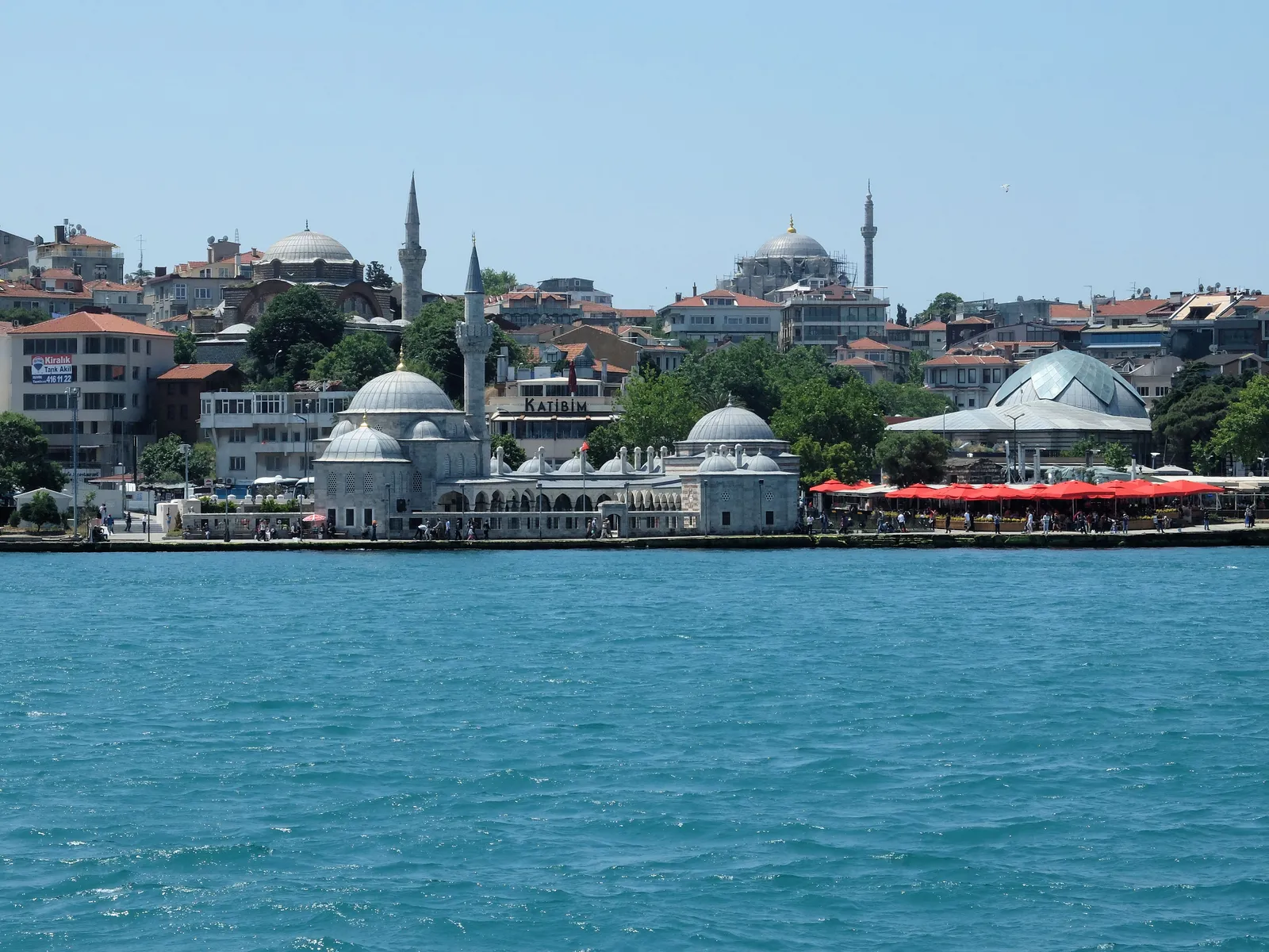 Şemsi Pasha Mosque on the Üsküdar shoreline seen from the water, a small domed mosque and its attached tomb standing right at the sea wall with a single minaret, red café parasols along the quay beside it