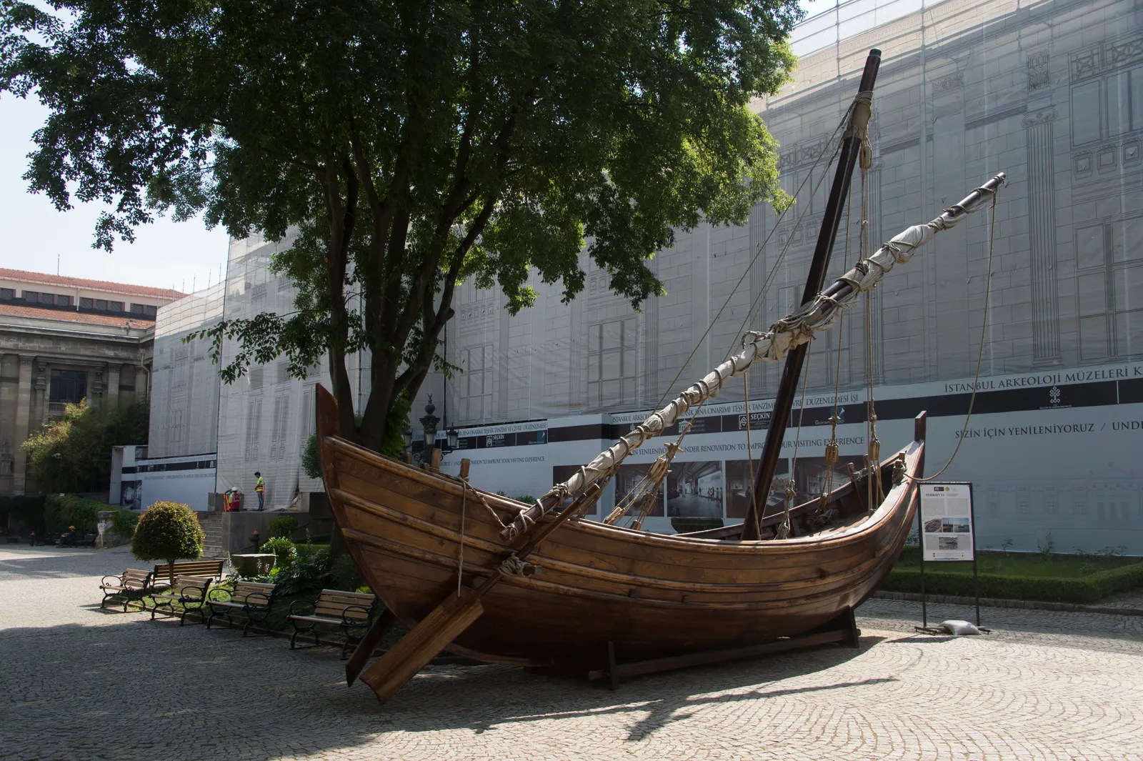 A full-size reconstruction of a small 9th-century Byzantine merchant ship standing in the garden of the Istanbul Archaeology Museums, its wooden hull and single mast set up under trees beside the museum building