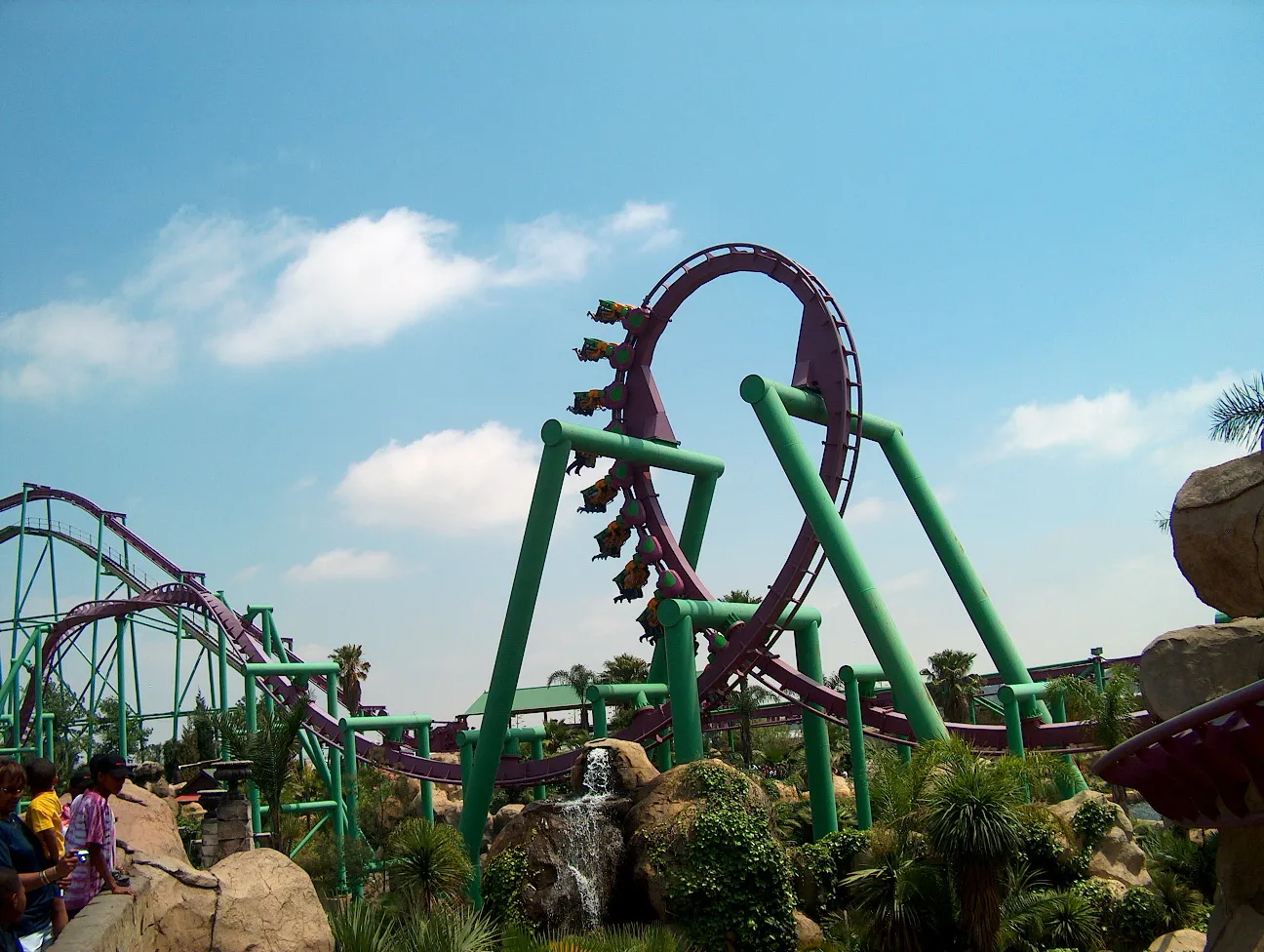 The Anaconda roller coaster at Gold Reef City in Johannesburg, a purple track on green supports curving through a vertical loop with a train of riders hanging below the rails, rocks and palms at ground level