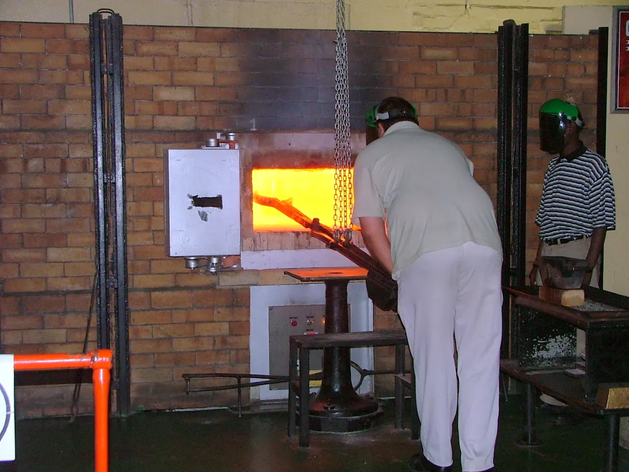 The gold pour at Gold Reef City in Johannesburg, a man in a green visor drawing a long-handled crucible out of the white-hot mouth of a brick arc furnace while a second man in a visor watches from behind a barrier
