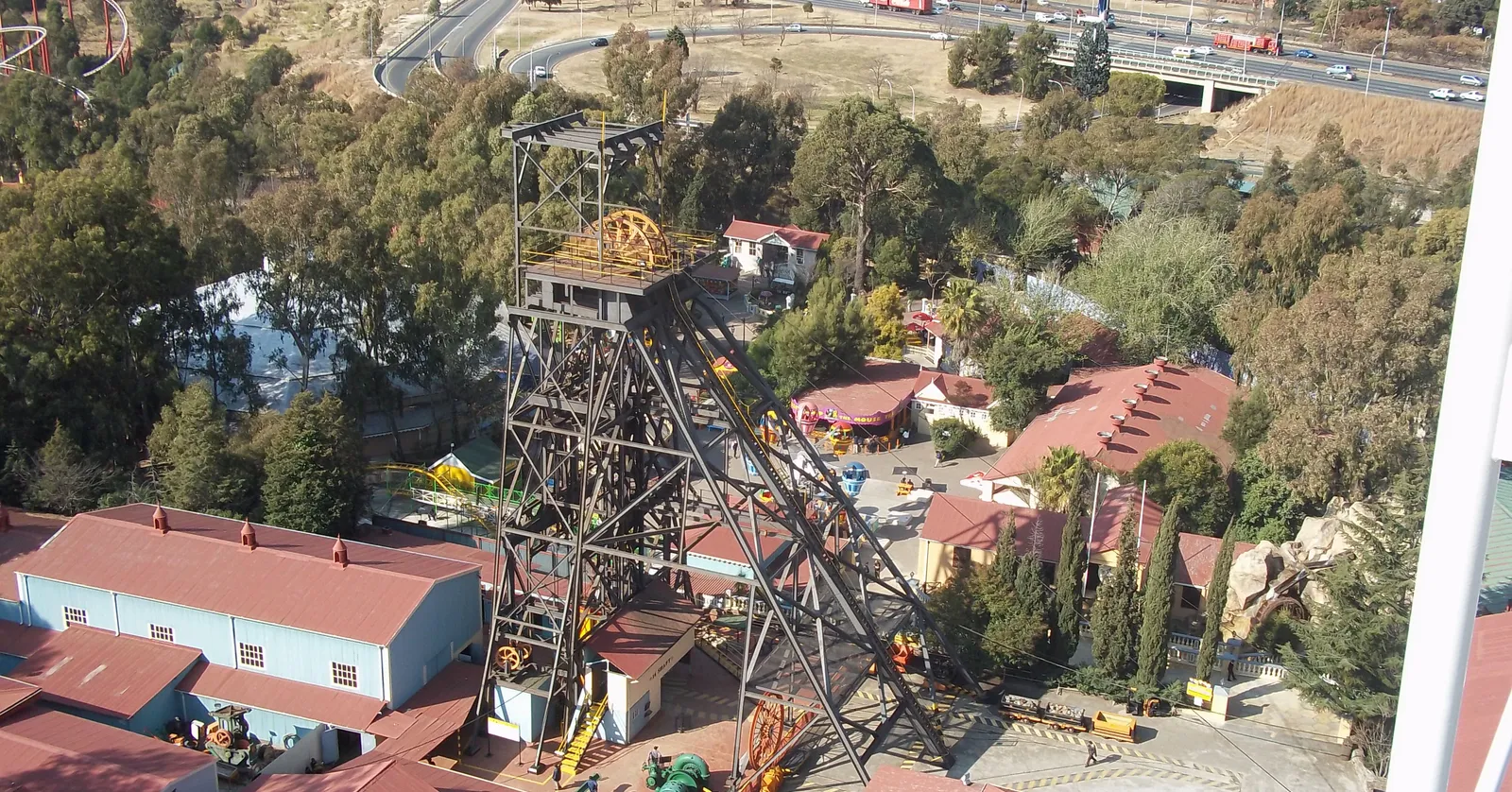 The steel mine headgear at Gold Reef City in Johannesburg seen from above, its yellow winding wheel at the top of a black lattice tower, standing over the red-roofed buildings and paved walkways of the park with a motorway beyond