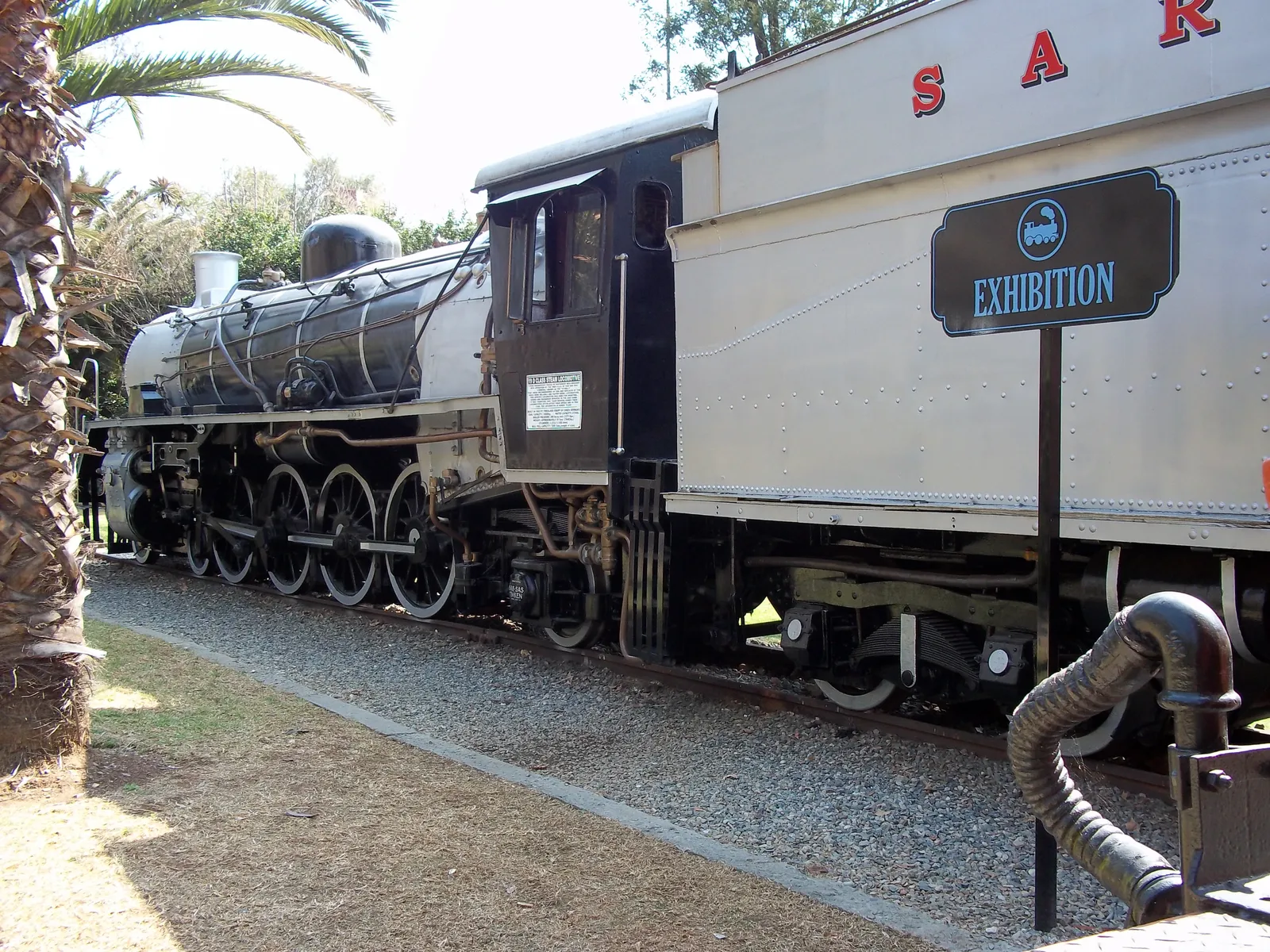 A black steam locomotive at Gold Reef City in Johannesburg coupled to a cream and maroon South African Railways coach lettered SAR, standing on a short length of track beside a gravel path and a palm tree