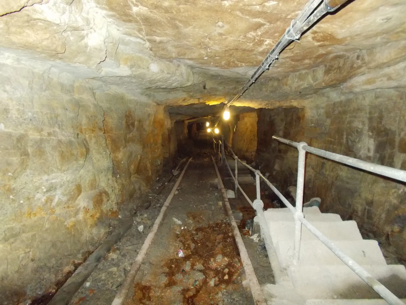 A tunnel underground at Gold Reef City in Johannesburg, rough pale rock walls and roof running away into the distance, a narrow rail track down the middle of the floor, a handrail and concrete steps on the right and a string of bulbs lighting the far end