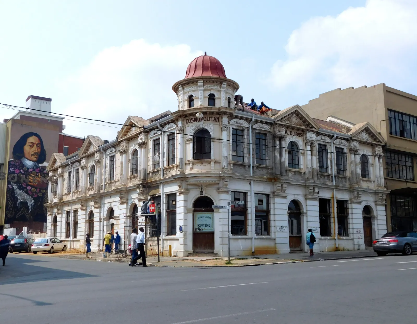 The Cosmopolitan on Commissioner Street in Johannesburg, a two-storey corner building with a red-domed turret, moulded plasterwork and arched windows, with a large painted portrait covering the side wall of the building next door