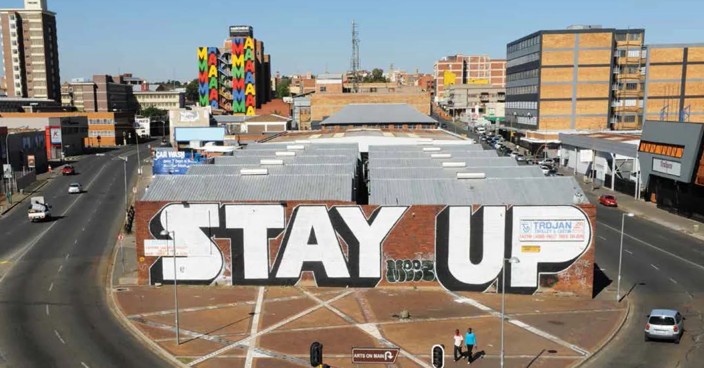 Warehouse roofs in Maboneng, Johannesburg, seen from above, with STAY UP painted in huge block letters across the brick end wall facing a paved street junction, and a tower block covered in coloured lettering standing behind it