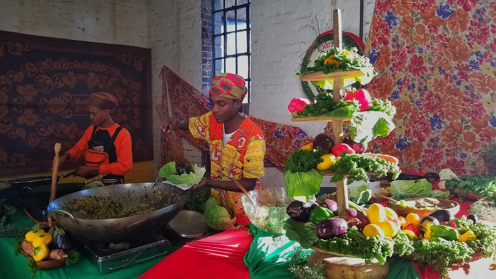 A produce stall at Market on Main in Johannesburg, two traders behind a tiered display of vegetables and a wide pan of cooked food, inside a brick warehouse hall with tall steel-framed windows