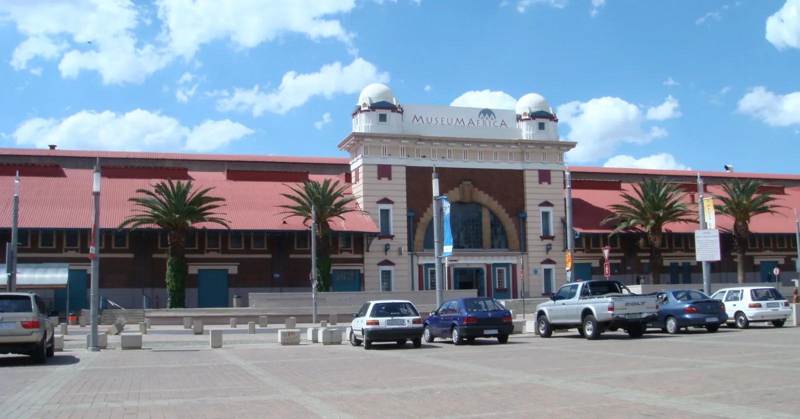 Museum Africa in Newtown, Johannesburg, a long red-roofed brick market shed with a pale central block carrying two small domes and the museum's name, palm trees and parked cars on the brick apron of Mary Fitzgerald Square in front