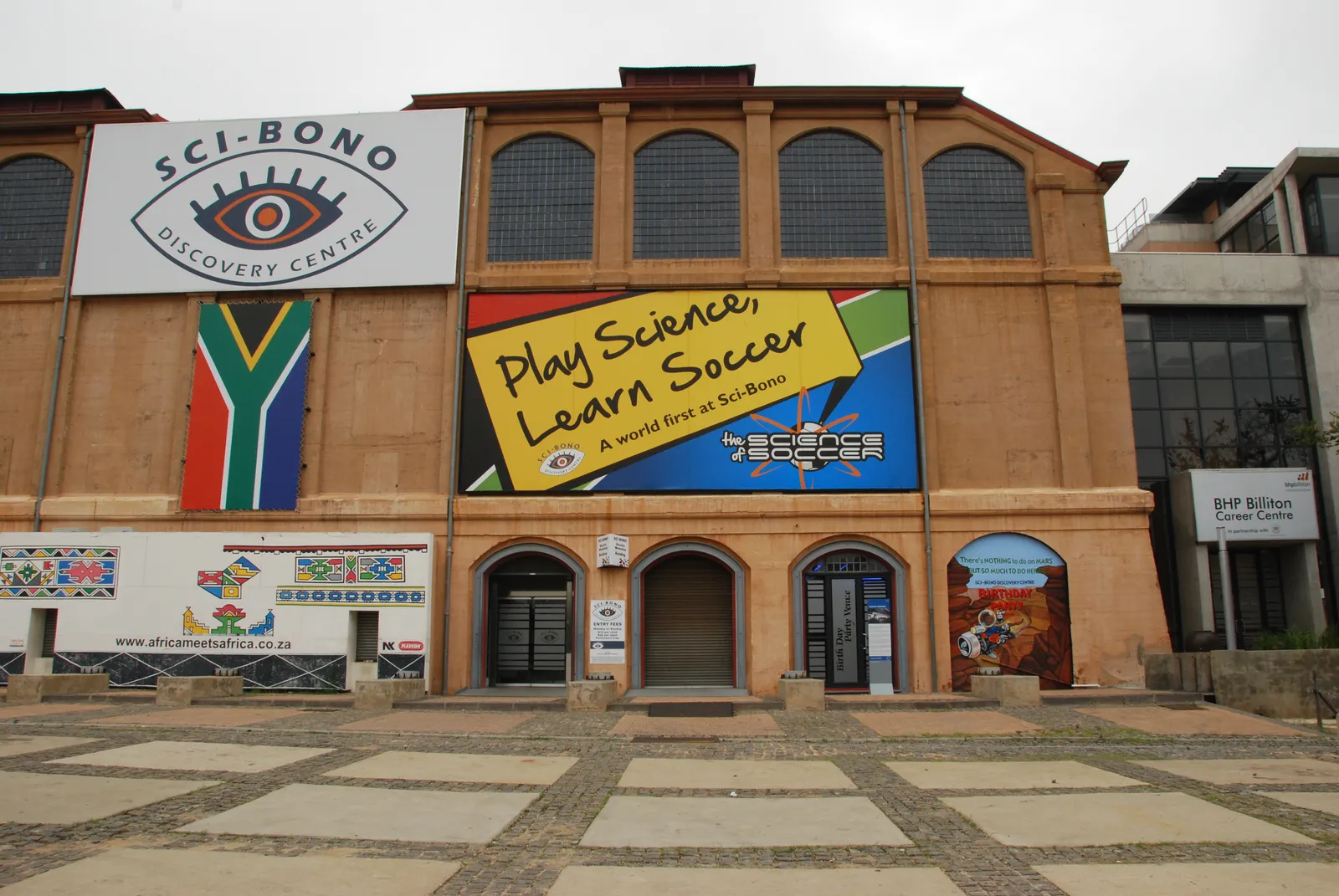 The Sci-Bono Discovery Centre in Newtown, Johannesburg, a long brick industrial hall with tall arched windows along its upper wall and a painted chequerboard forecourt in front of the doors
