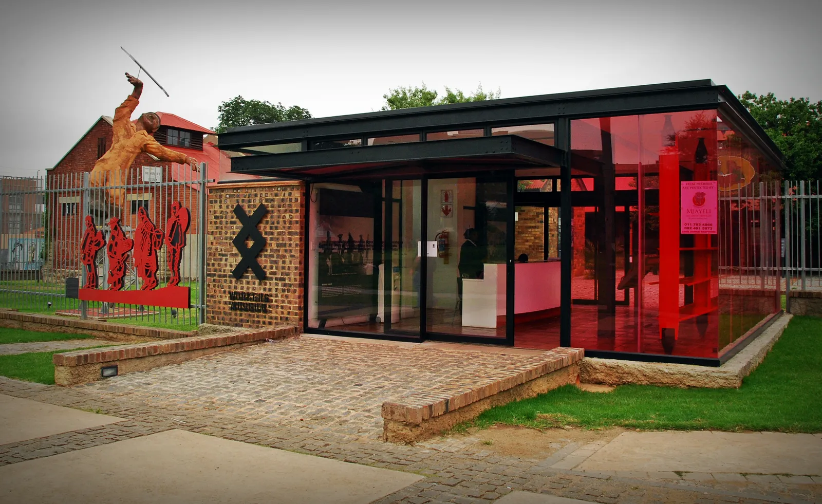 The glass and red-steel entrance pavilion of the Workers Museum in Newtown, Johannesburg, standing on a brick apron in front of the low red-roofed brick compound buildings behind it