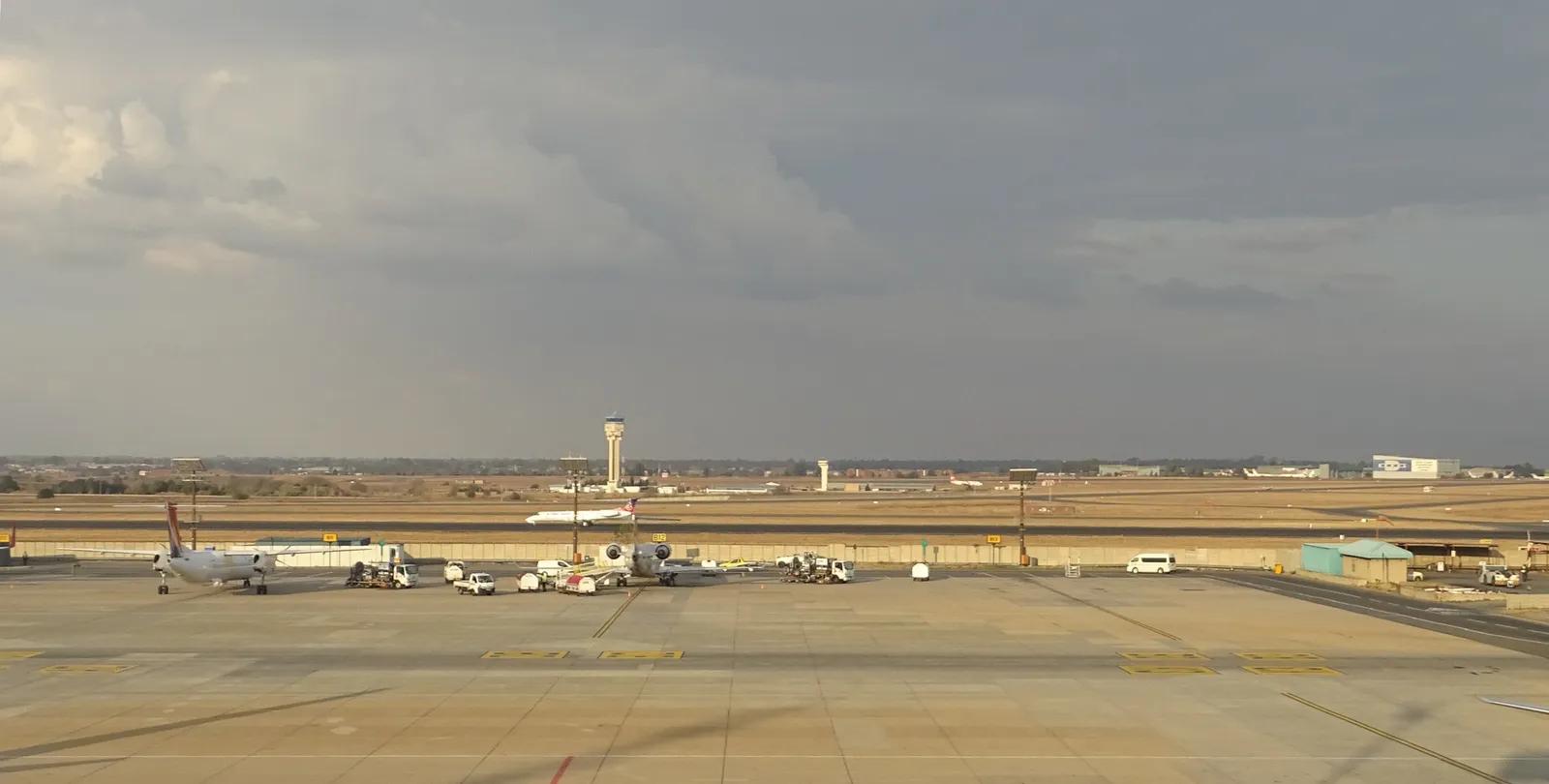 The apron at O. R. Tambo International Airport in Johannesburg under a grey sky, aircraft parked at stands with ground vehicles around them and the control tower standing beyond the taxiways