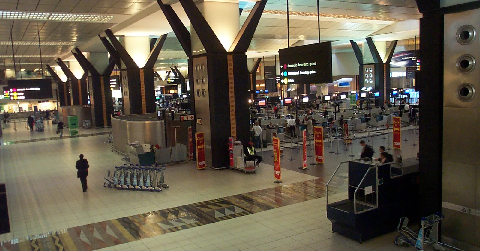 A check-in hall at O. R. Tambo International Airport in Johannesburg seen from the level above, rows of counters under angled black columns, luggage trolleys lined up on a patterned stone floor and overhead signs for domestic and international boarding gates