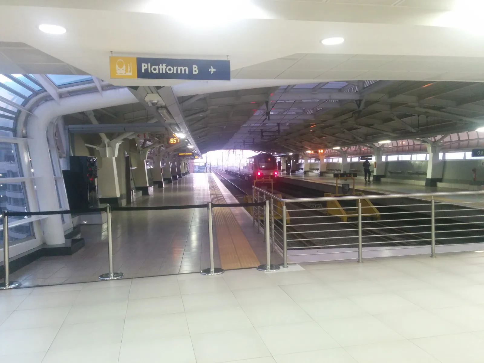 The platform level of the train station inside O. R. Tambo International Airport in Johannesburg, a vaulted white roof over an empty platform with a sign reading Platform B and a train pulling away down the track