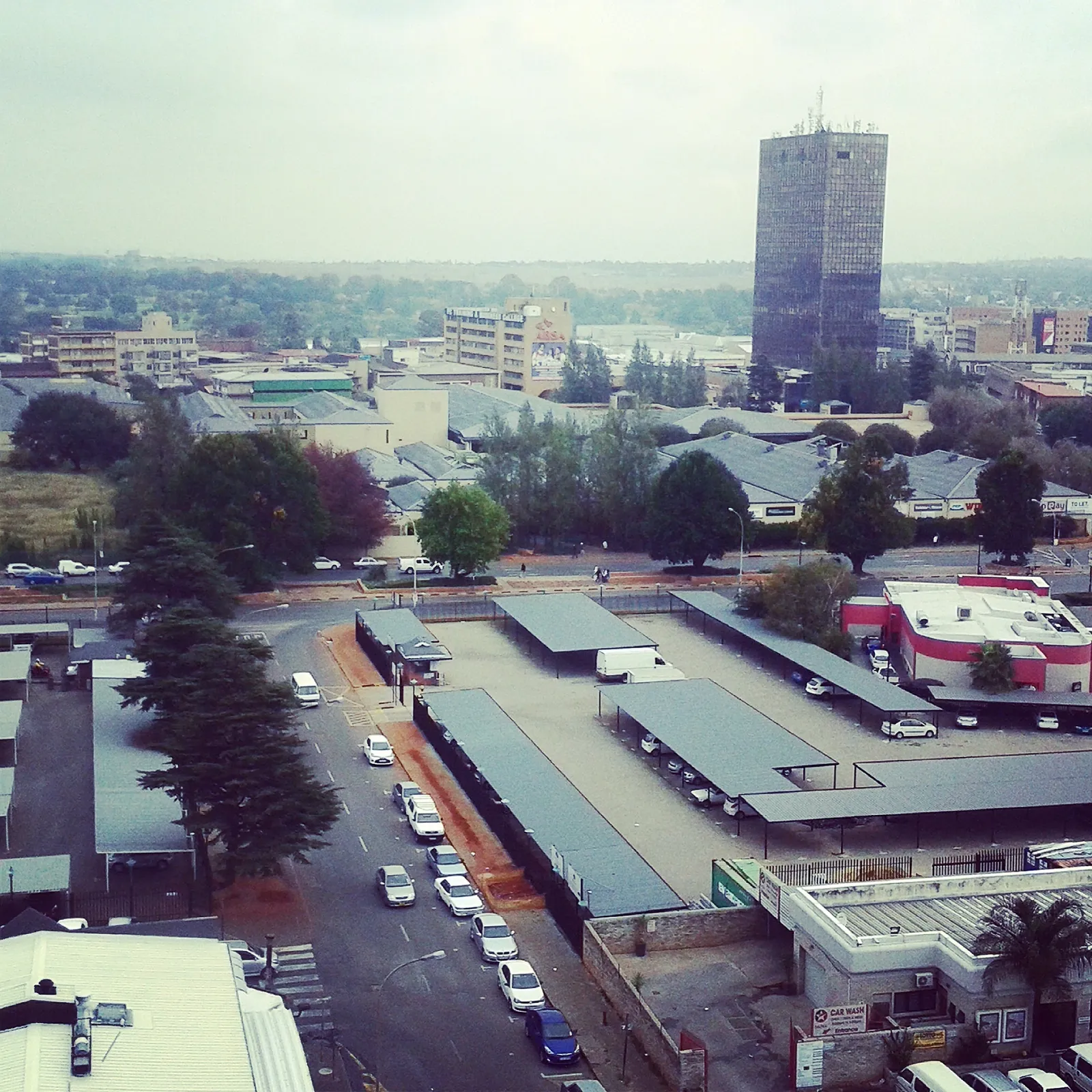 The centre of Kempton Park from above, a dark high-rise block standing over low shopfronts and warehouse roofs, with cars parked along a wide street lined with pines