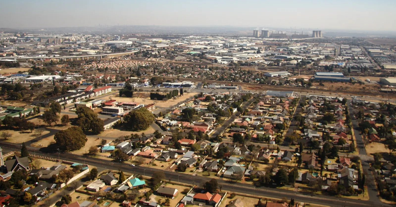 Rhodesfield in Kempton Park seen from the air, streets of single-storey houses with pitched red and green roofs laid out between wide tarred roads, with warehouse roofs and power station cooling towers along the horizon