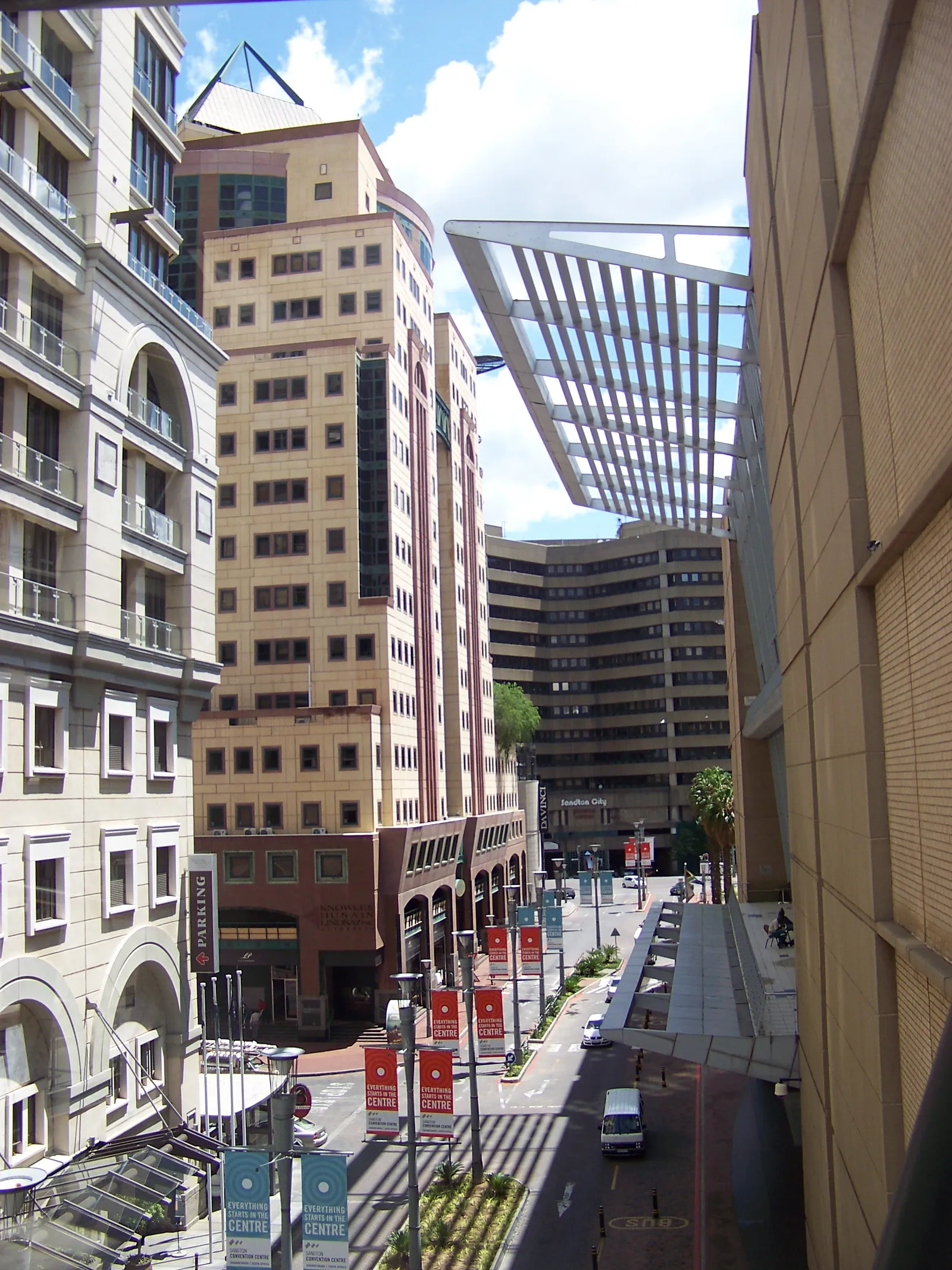 Maude Street in Sandton, Johannesburg, seen from above: office and apartment towers rising on both sides of a narrow street lined with banners, with a glass canopy over the pavement on the right