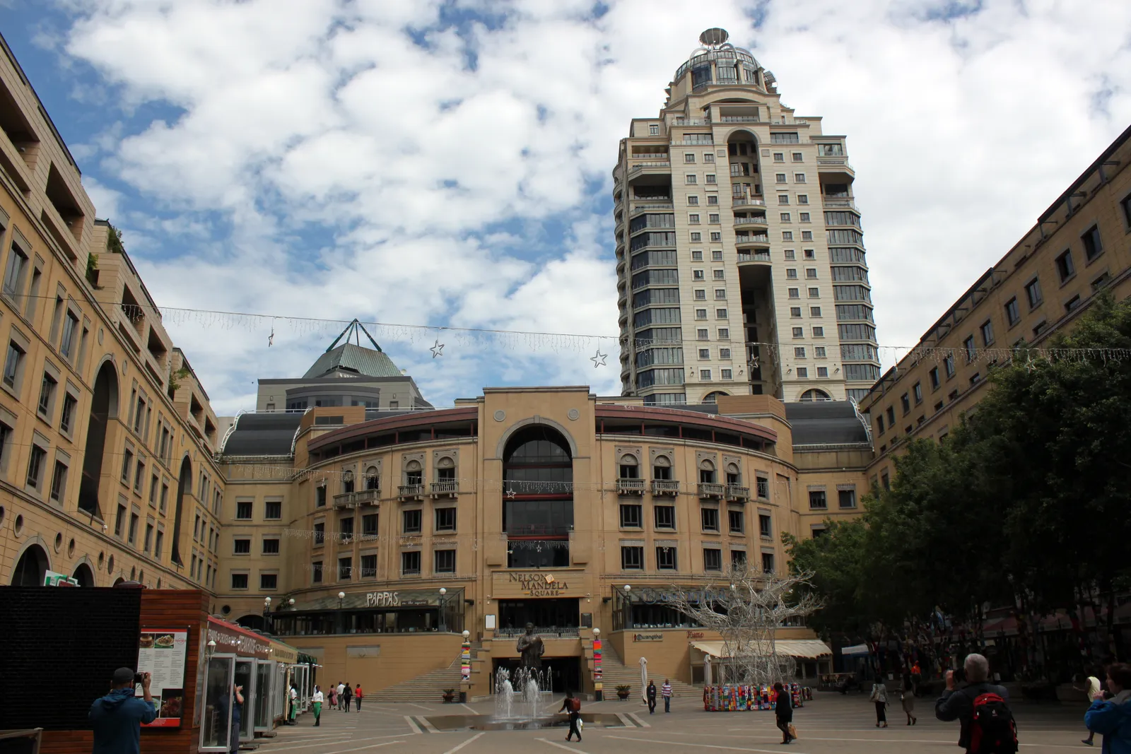 Nelson Mandela Square in Sandton, Johannesburg, an open paved piazza with a fountain in the middle, arcaded stone-coloured buildings on three sides and a tall residential tower with a crowned top rising behind