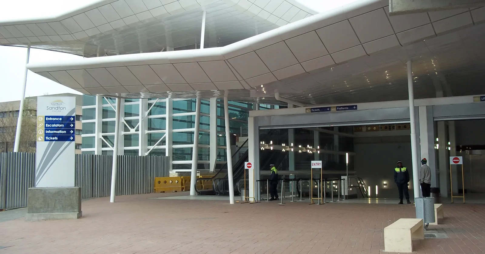 The street entrance to Sandton station in Johannesburg, a curved white canopy on slender columns over a paved forecourt, with glass doors, ticket signage and staff in high-visibility vests underneath