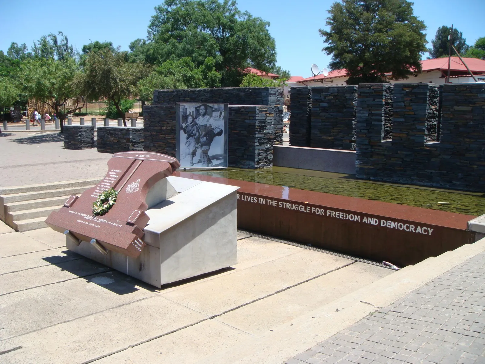 The Hector Pieterson Memorial in Orlando West, Soweto, a brick and stone plaza with a shallow water channel, an enlarged photograph mounted on a dark stone wall and an inscription reading LIVES IN THE STRUGGLE FOR FREEDOM AND DEMOCRACY