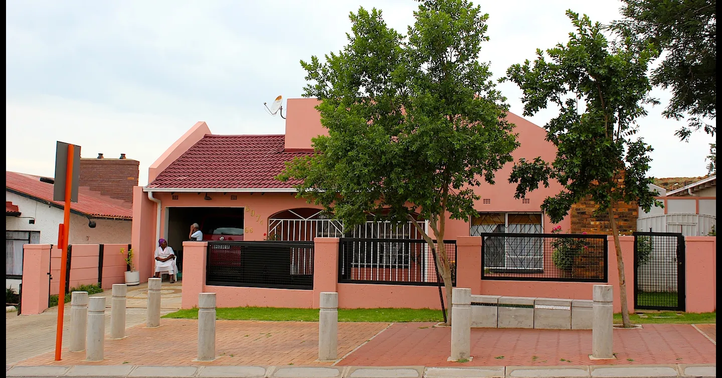 A salmon-pink house behind a low wall and black railings on Vilakazi Street in Soweto, Johannesburg, with a resident sitting in the shade of the carport and concrete bollards lining the paved pavement in front