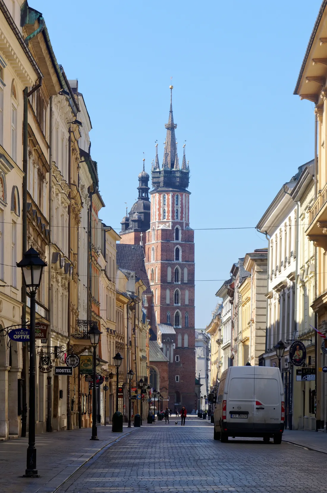 Floriańska Street in Kraków looking south between rows of pastel townhouses and hanging shop signs, with the two brick towers of St Mary's Basilica closing off the far end of the cobbled street