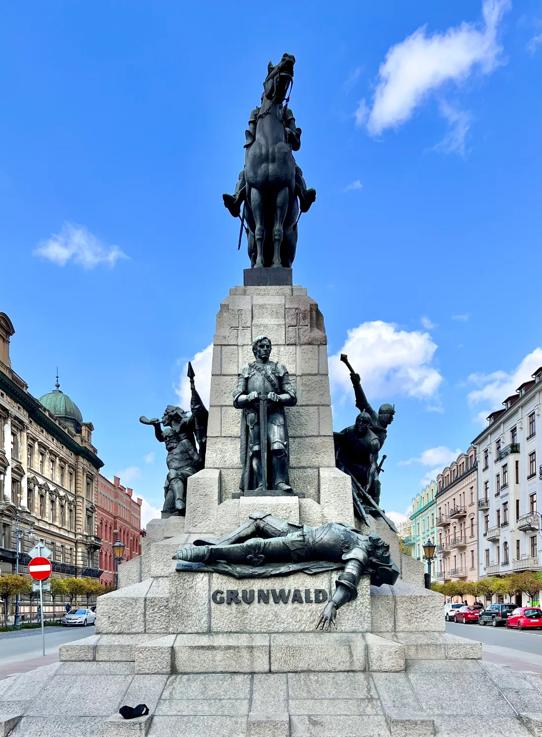The Grunwald Monument in Kraków, a bronze king on horseback on top of a tall stone plinth lettered GRUNWALD, with armoured figures around its base and a fallen knight lying across the front step