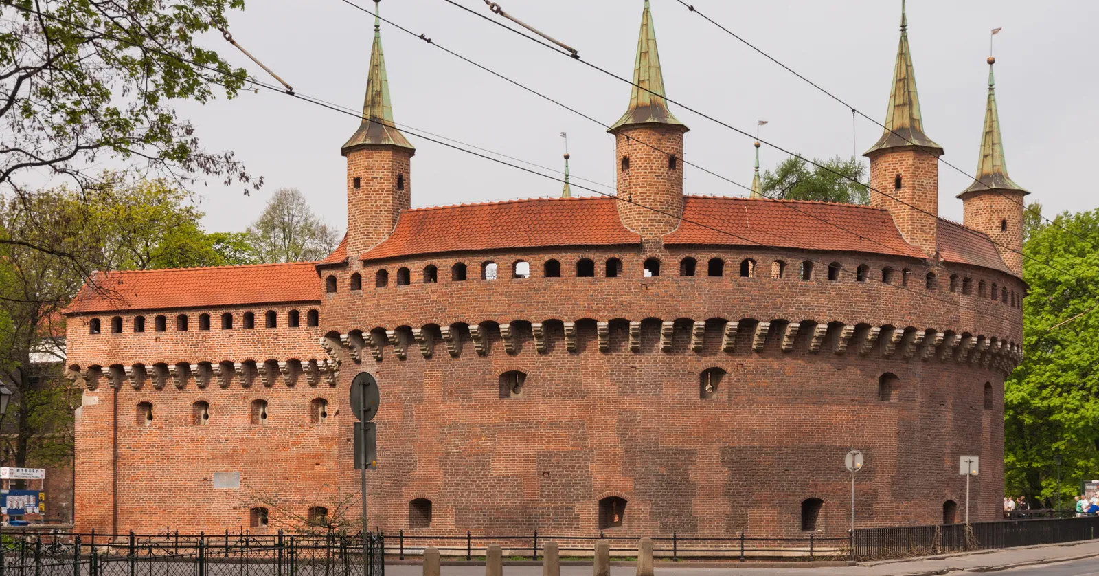 The Kraków Barbican, a squat circular fort of red brick ringed by a gallery of arrowslits, with four pointed turrets rising from its roofline and trees of the surrounding park on either side