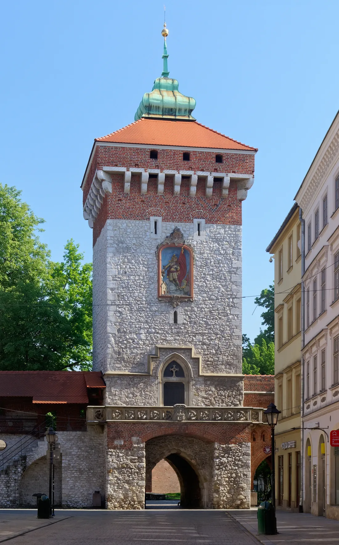 St Florian's Gate in Kraków seen head on, a square tower of pale stone below and red brick above under a tiled roof and a green copper cupola, with a painted relief panel set into the wall over its archway