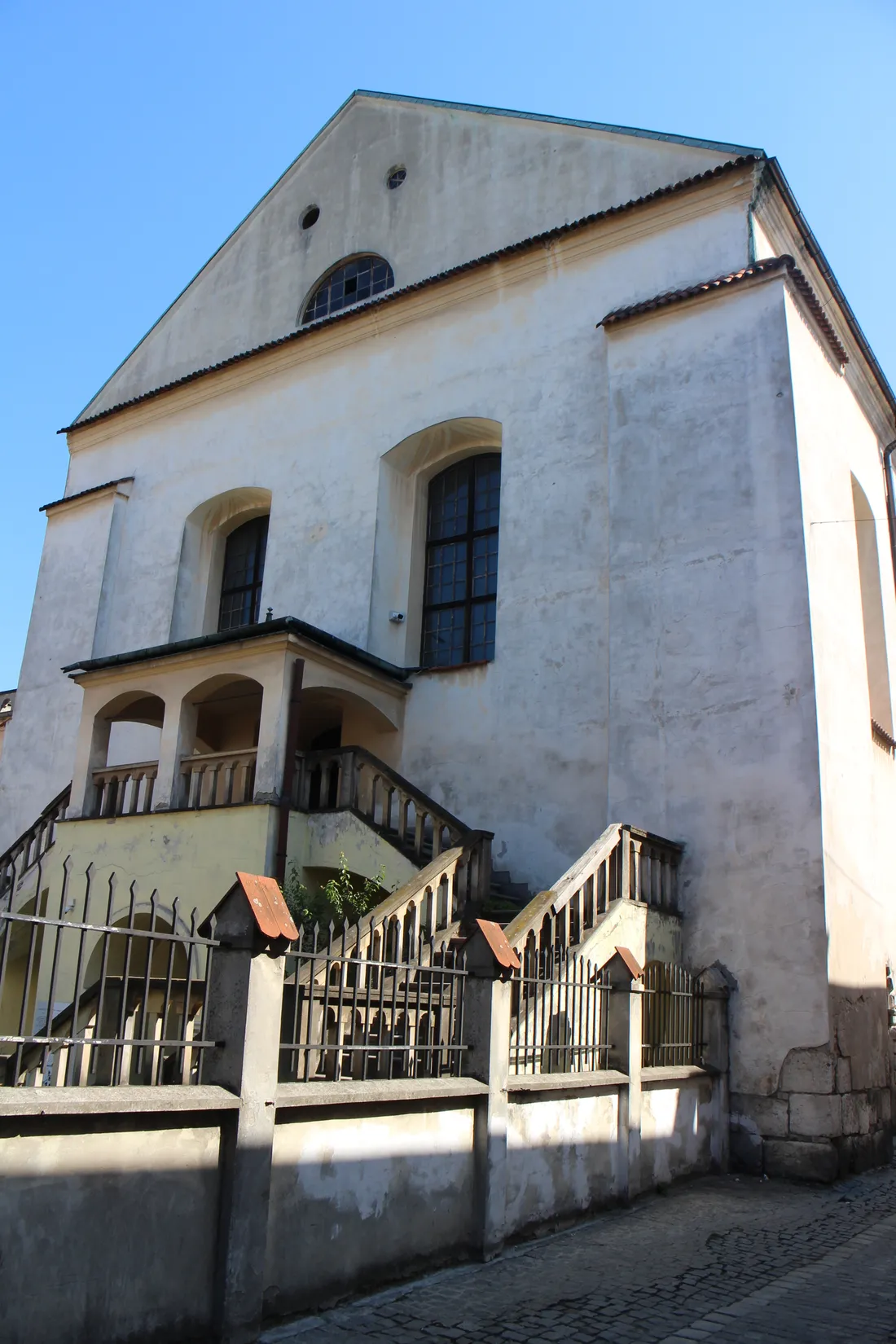 The Isaac Synagogue in Kraków, a tall whitewashed hall with a plain gable and two arched windows, an outdoor stone staircase running up to a covered porch along its side, seen from the cobbled street below