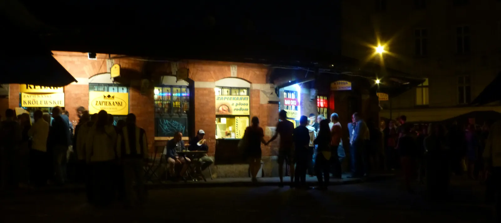 The round brick market hall on Plac Nowy in Kraków at night, lit food hatches cut around its outside wall with signs above them and a crowd standing and eating in the dark square