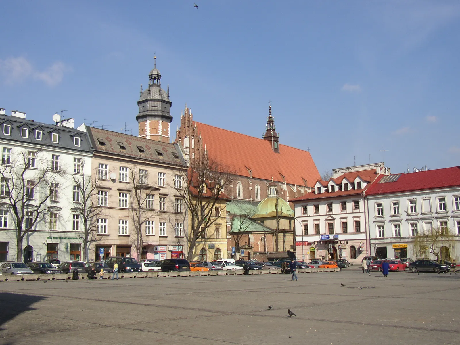 Plac Wolnica in Kraków, a wide open square with parked cars along the far side, the neo-Renaissance town hall tower on the left and the tall red roof of the Corpus Christi Basilica behind the townhouses