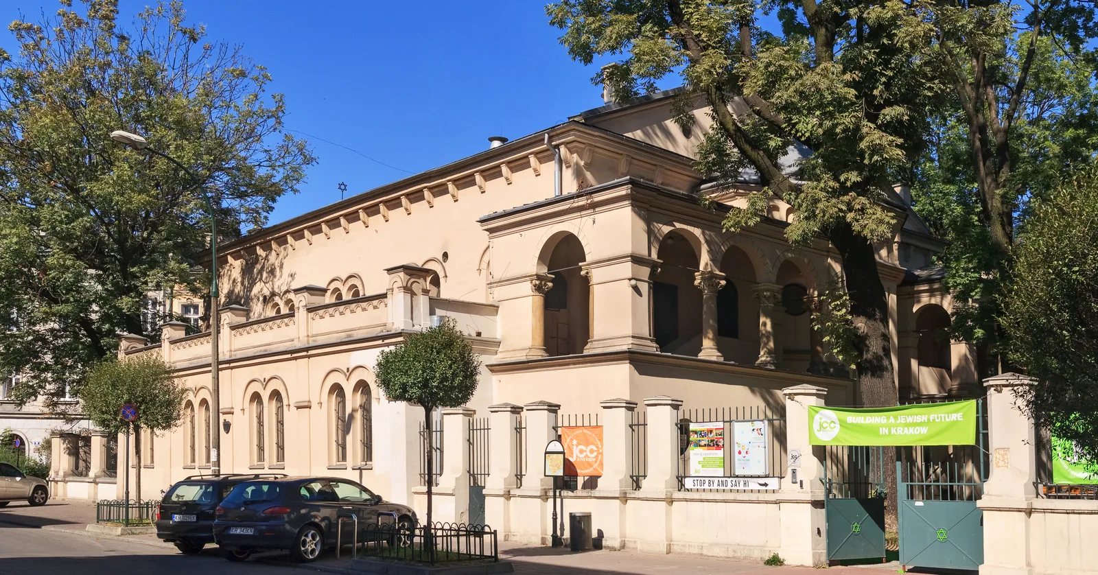 The Tempel Synagogue in Kraków's Kazimierz seen from the street corner, a long sand-coloured 19th-century building with rows of round Moorish arched windows, behind a low wall and railings with trees on either side