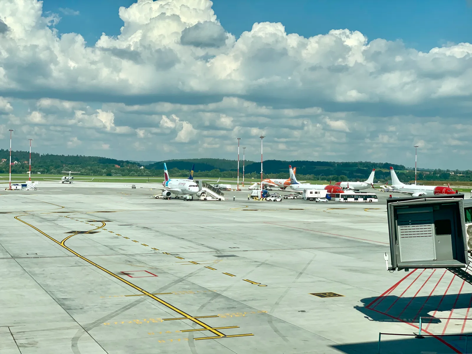 The apron at Kraków Airport seen from inside the terminal, aircraft parked in a row with boarding stairs and service vehicles around them, yellow taxiway markings across the concrete and wooded hills on the horizon