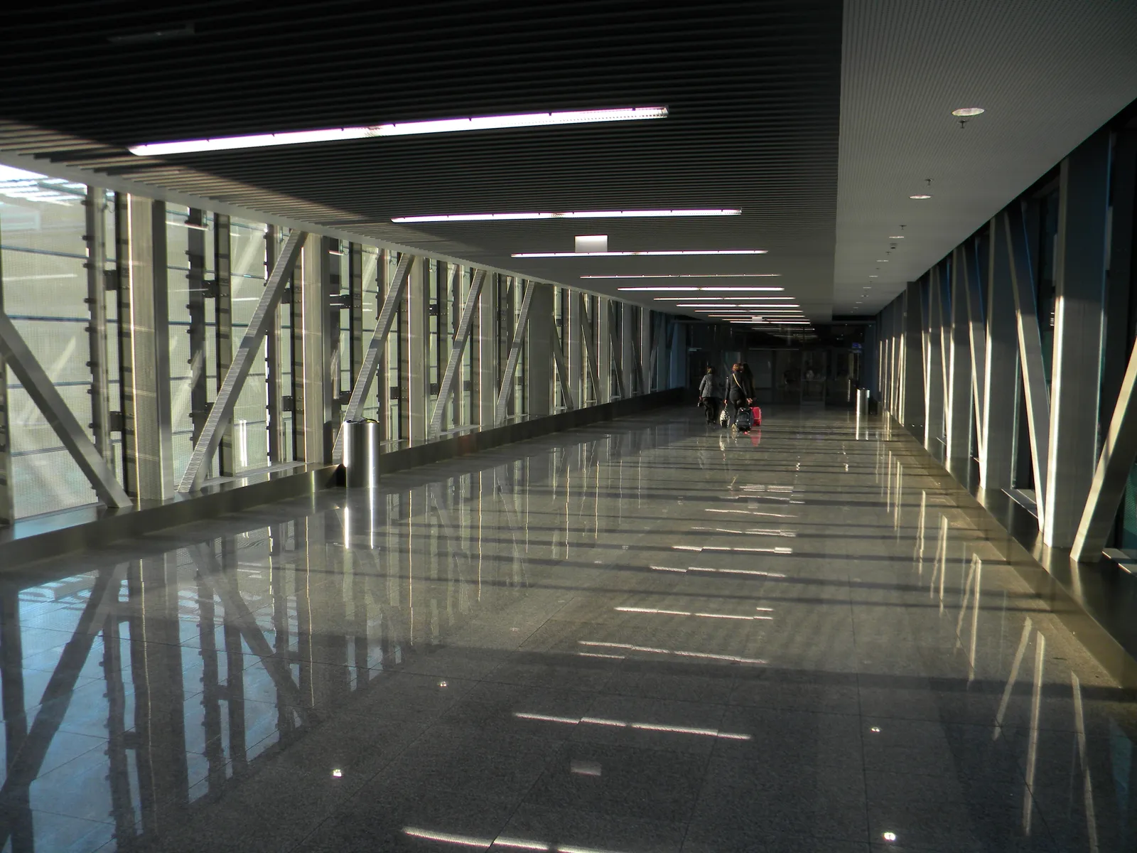 The covered footbridge between Kraków Airport's terminal and its railway platform, a long glazed corridor with a polished dark floor and steel bracing down one side, two travellers with wheeled bags at the far end
