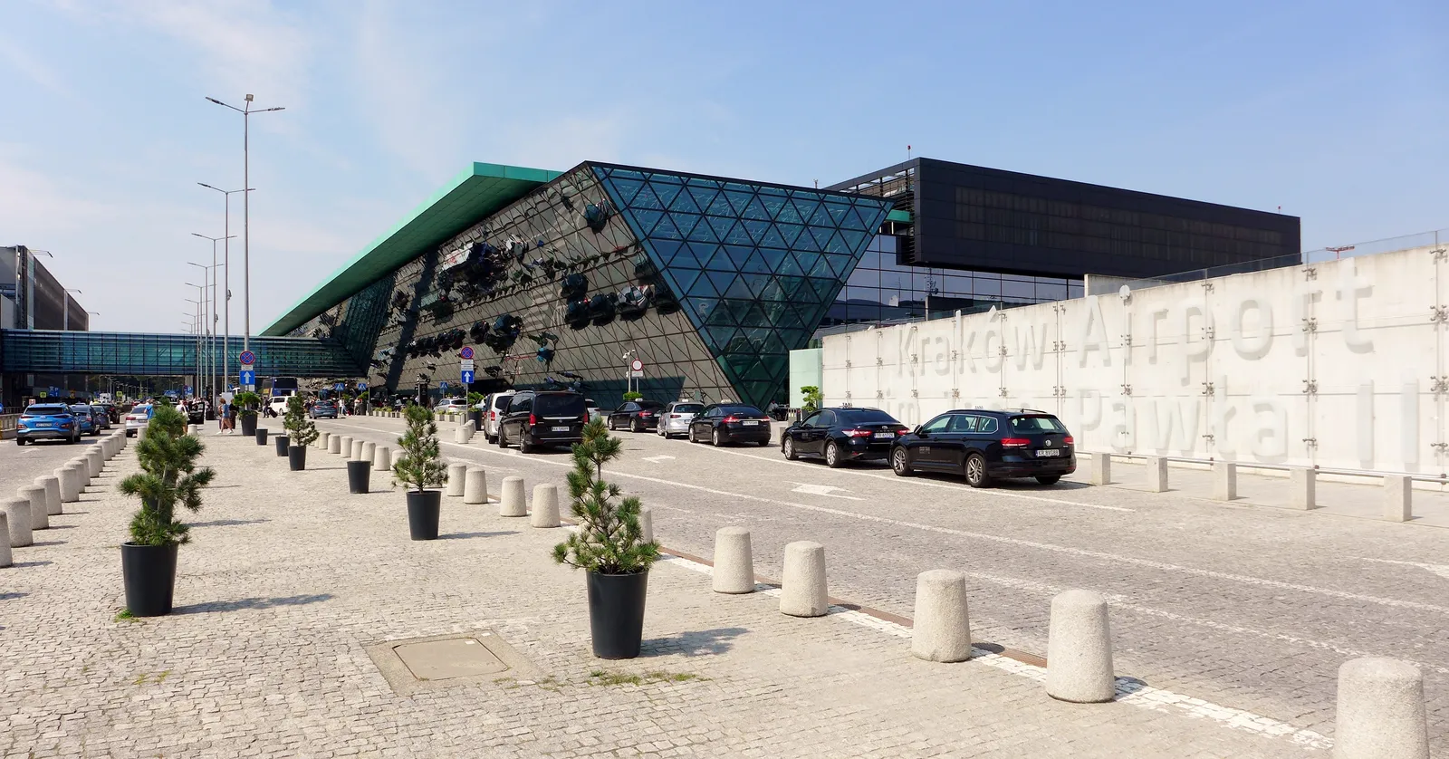 The front of Kraków Airport's passenger terminal, a long dark building with a faceted green and glass wedge over the entrance and the airport's name in tall letters on a wall beside the drop-off road