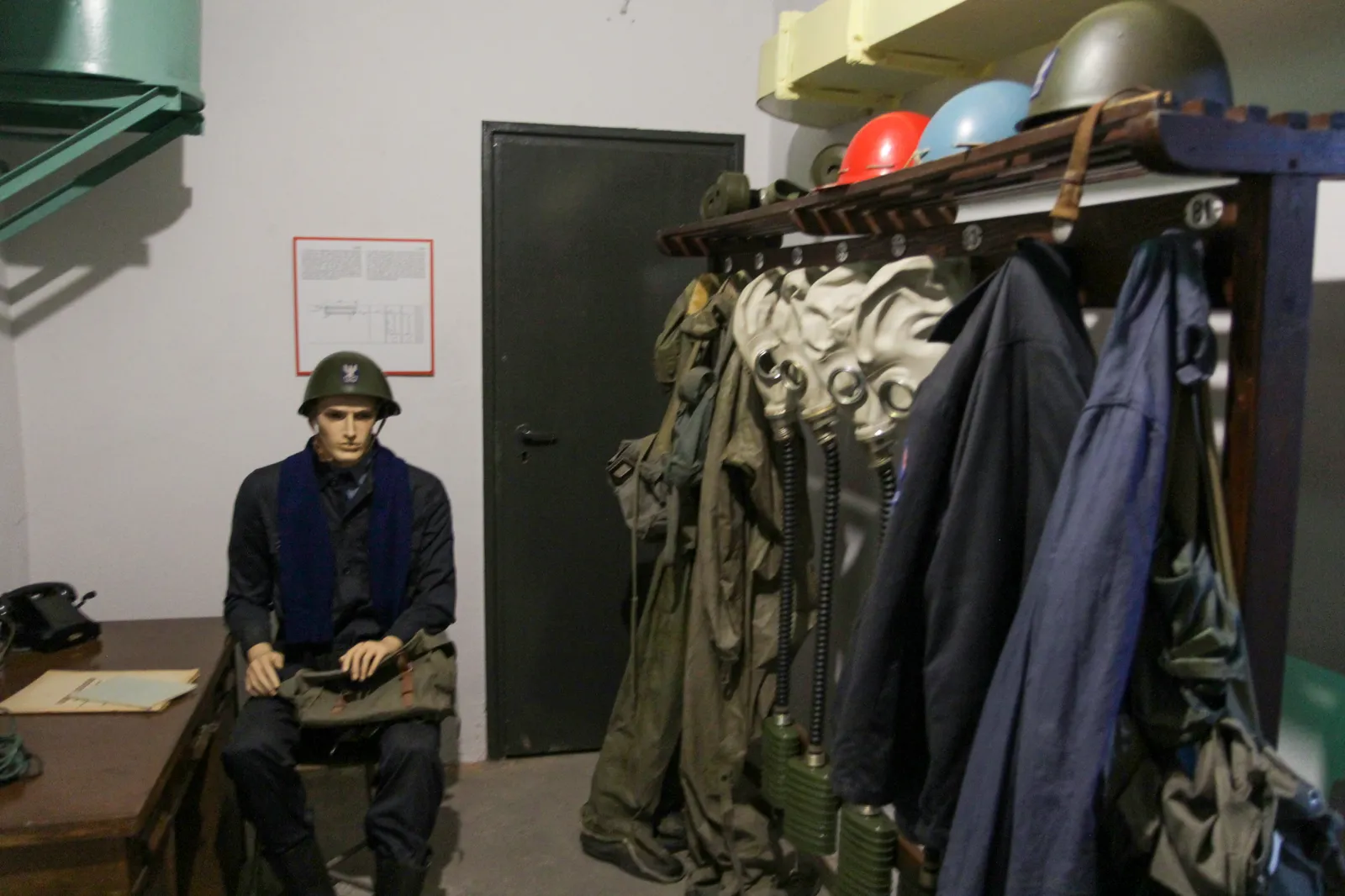 Inside a Cold War shelter under Nowa Huta, a low concrete room with a steel blast door, racks of civil defence coats, gas masks and helmets along one wall and a mannequin in a helmet sitting at a desk