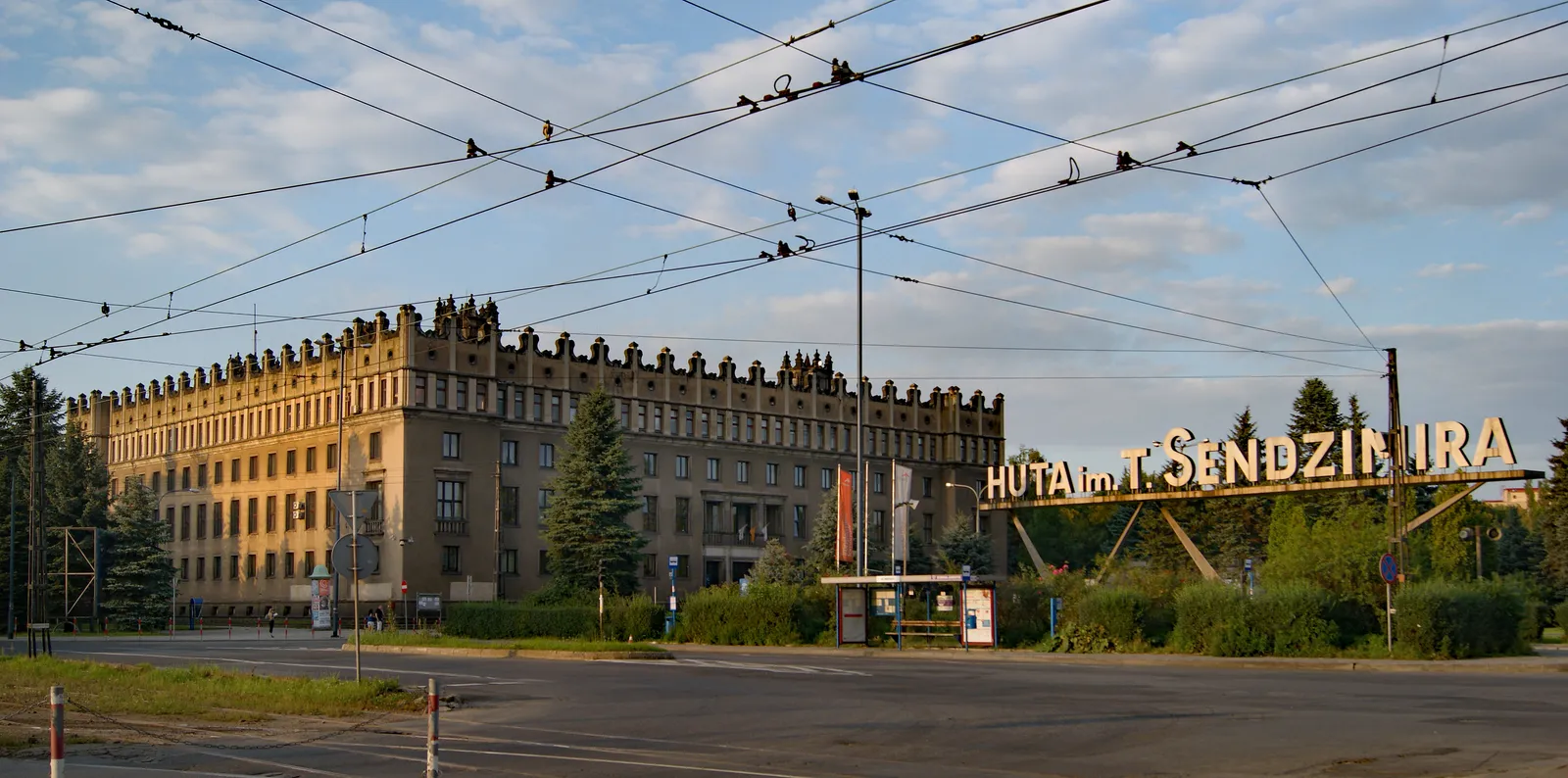 One of the two Renaissance-styled administration blocks at the Nowa Huta steelworks gate in Kraków, a long stone building with an ornamental parapet along its roofline, tram wires overhead and the works' name on a sign to the right