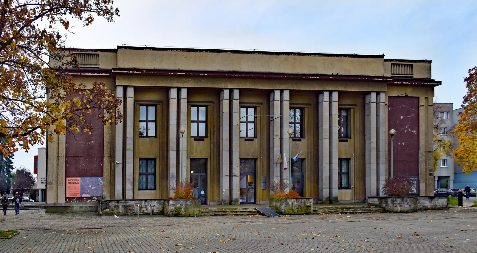 The former Światowid cinema in Nowa Huta, a symmetrical 1950s building with a row of tall square columns across its front and dark red panels at either end, standing on an empty paved forecourt