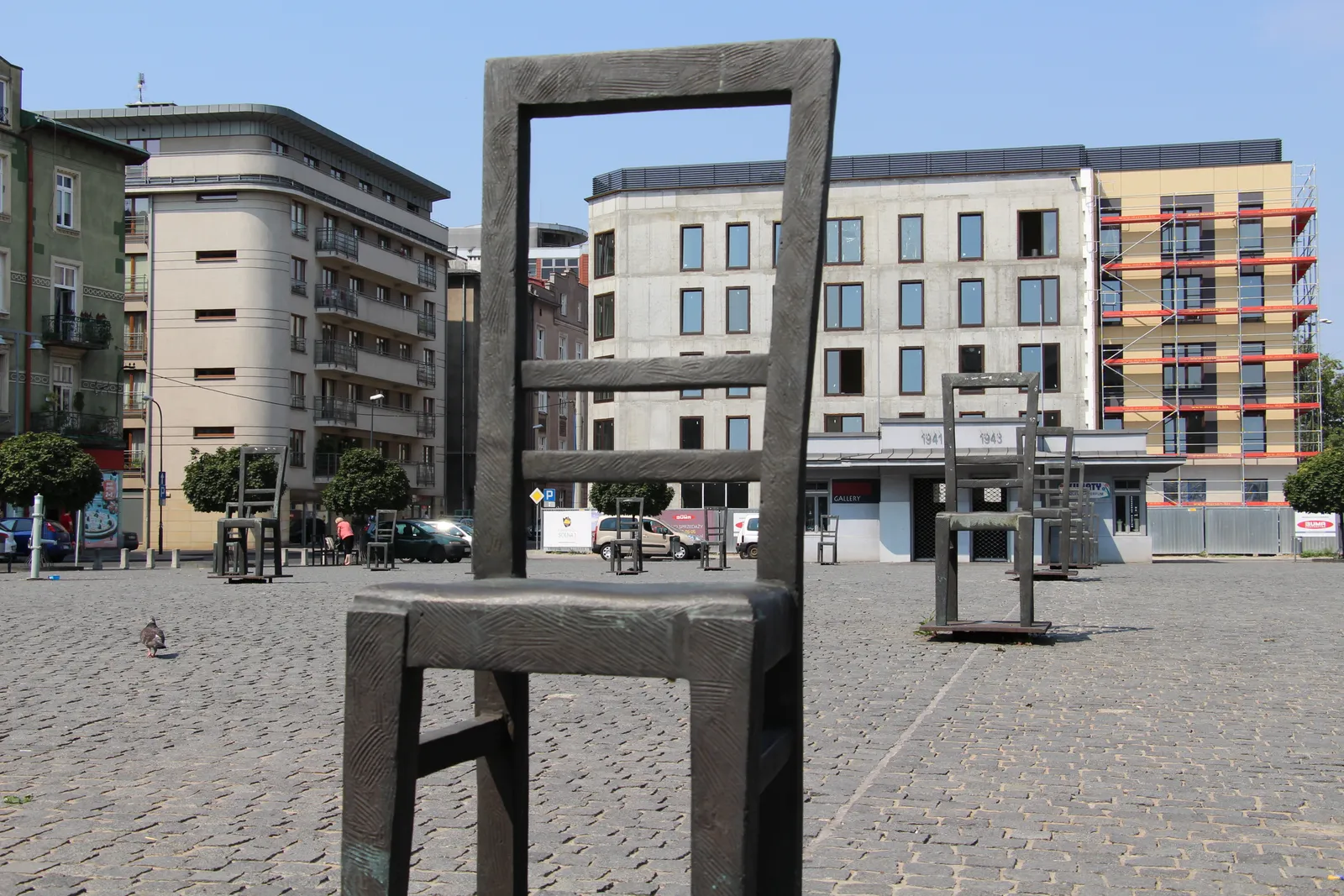 An oversized steel chair standing alone on the cobbles of Ghetto Heroes Square in Kraków, with more chairs spaced across the empty square behind it and apartment blocks along the far side