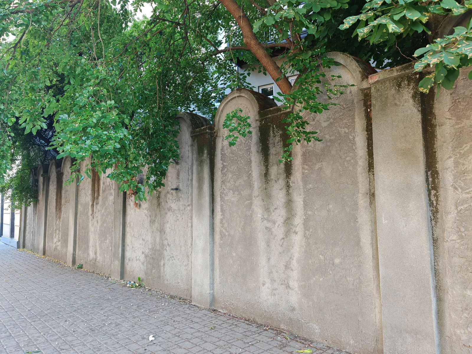 A surviving stretch of the Kraków ghetto wall on Lwowska Street, a rendered concrete wall whose panels are finished with rounded tops in the shape of Jewish gravestones, running along a paved pavement under trees