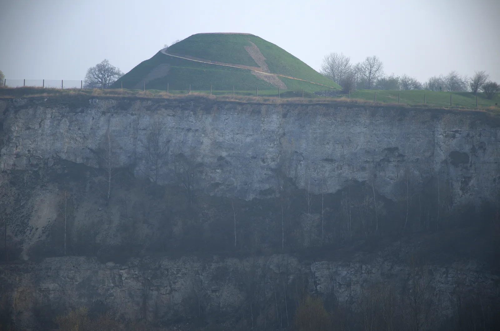 The grass-covered cone of Krakus Mound in Kraków rising behind the sheer grey rock face of the Liban quarry, with a line of bare trees along the cliff edge between them