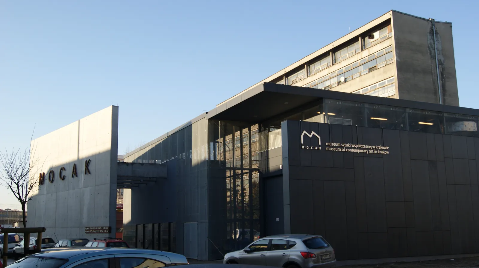 The MOCAK contemporary art museum in Kraków, a low dark grey building with sawtooth roofs and a glazed entrance bay, its name in large pale letters on the wall at the left