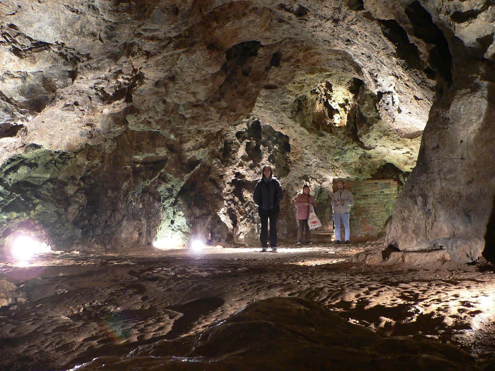 Inside the Dragon's Den under Wawel Hill in Kraków, a lit limestone chamber with a rough vaulted ceiling and a damp rock floor, three visitors walking through it for scale