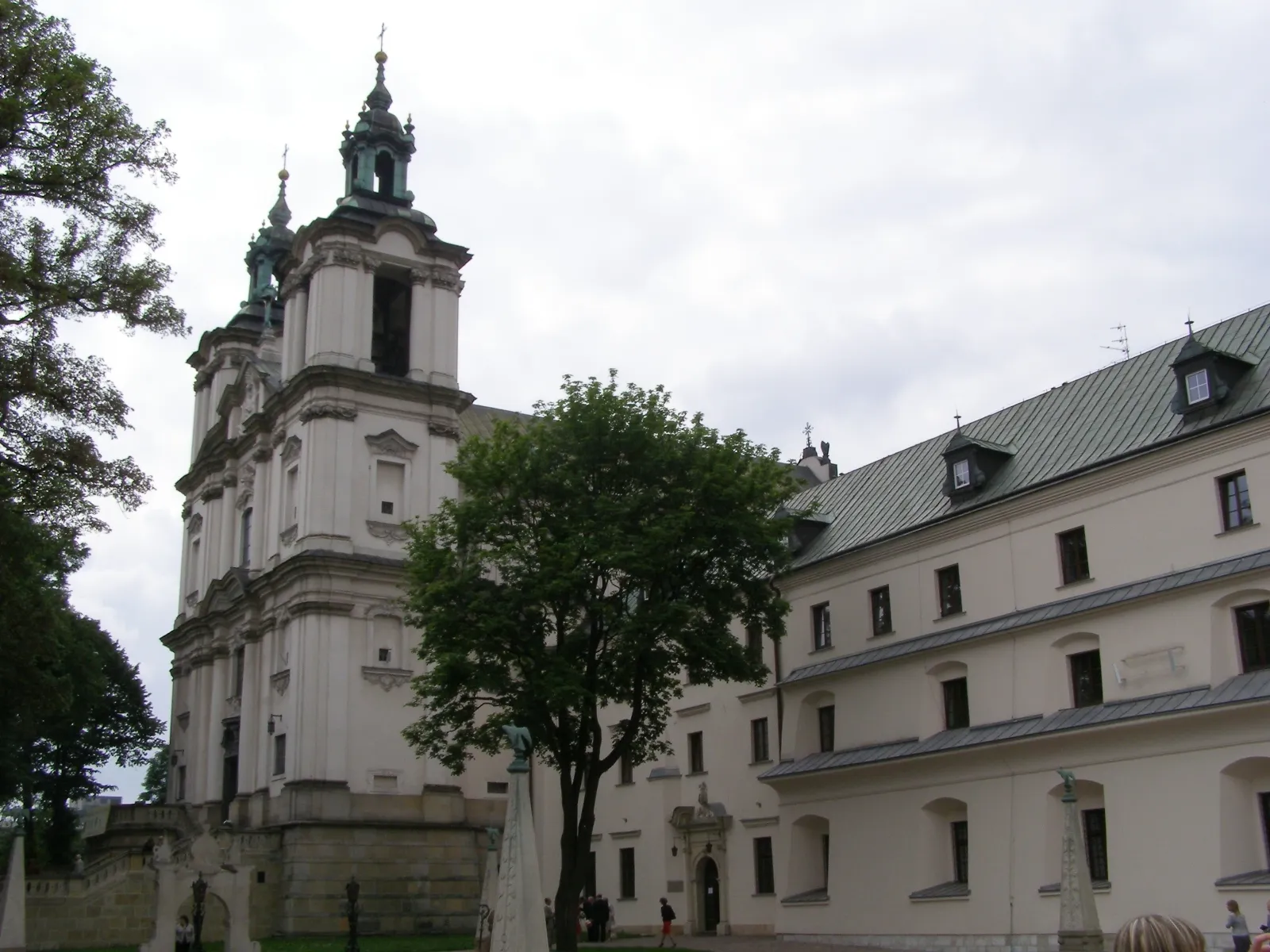 The white Baroque church at Skałka in Kraków, twin towers with dark copper cupolas above a columned front, with the long green-roofed monastery wing running back from it and a tree in the courtyard