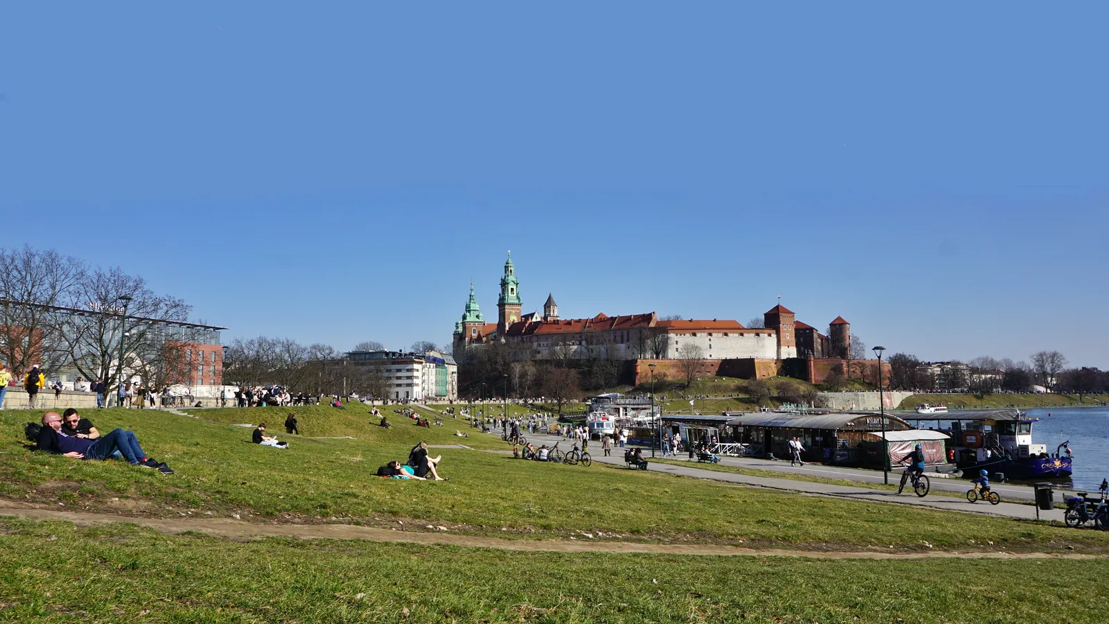 The grassy Vistula embankment in Kraków on a clear day, people sitting on the slope and walking the paved path below, with moored barges along the water and Wawel Hill's towers and red roofs rising behind